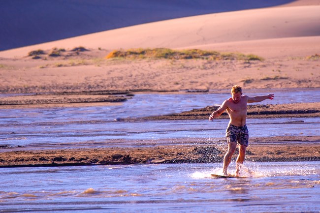 A skimboarder glides on a wide, shallow creek flowing at the base of dunes