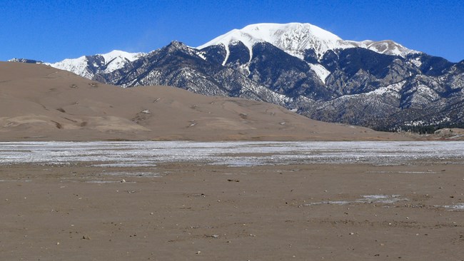 A dry sandy creek bed with partial snow cover, with dunes and a snow-covered mountain in the background