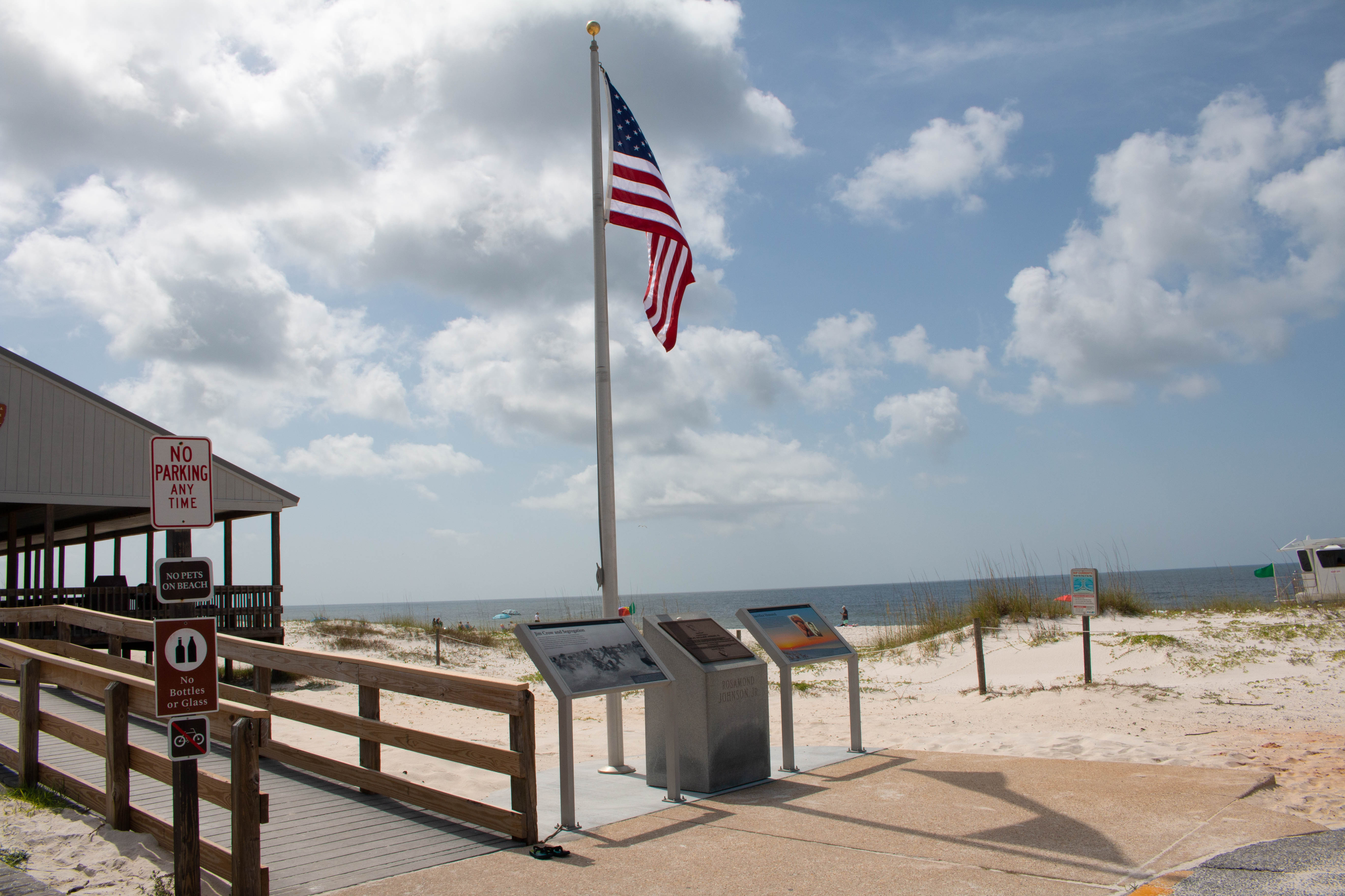 Beach pavilion on the left, two memorial plaques and an American flag in the center. White sand beach, turquoise water, and blue sky in the background.