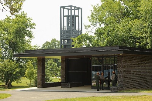 Three park ranger stand at the entrance of the new restroom facility at the U.S. Marine Corps Memorial. The restroom is one story tall, constructed from brick and glass with a flat, black roof. The building sits among trees and the Netherlands Carillon.