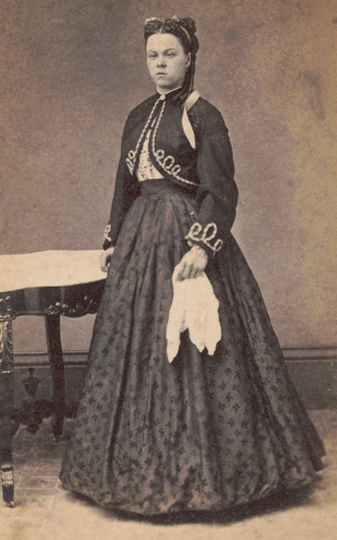 A young woman with long dark hair pulled back, wearing a dark long-sleeved jacket with white trim and long patterned skirt, holds a white handkerchief as she poses beside a low, ornate table.