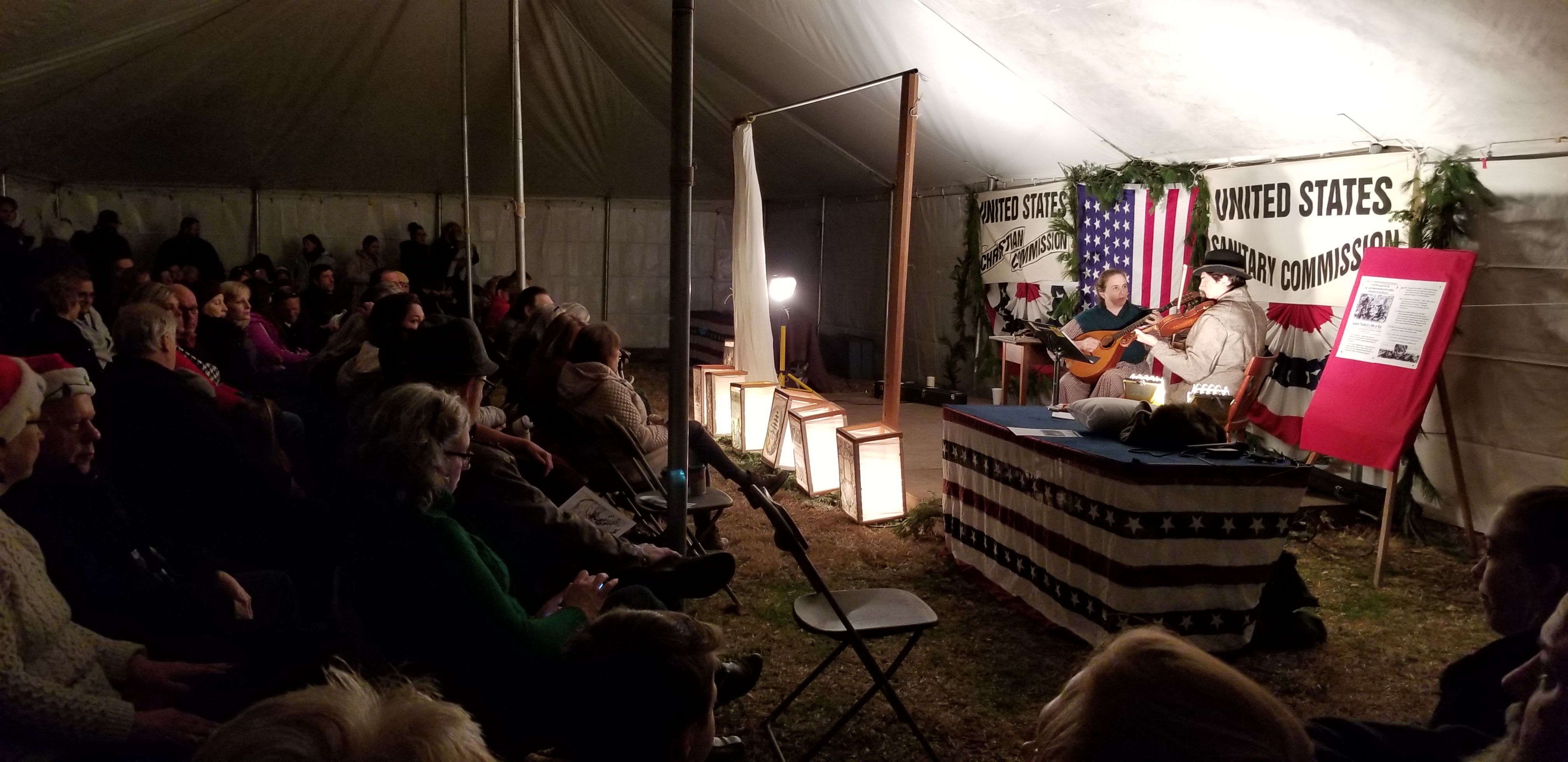 A crowd sits in a tent watching two musicians play stringed instruments