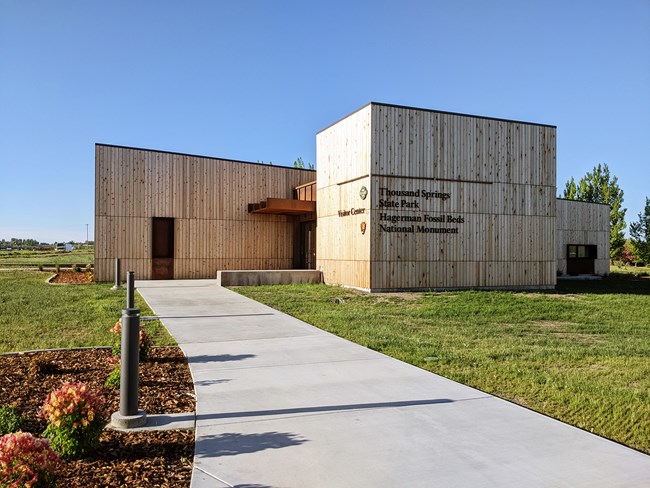 Exterior of the visitor center, a boxy building with wood plank exterior, landscaped lawn, and a paved sidewalk leading to the front doors.