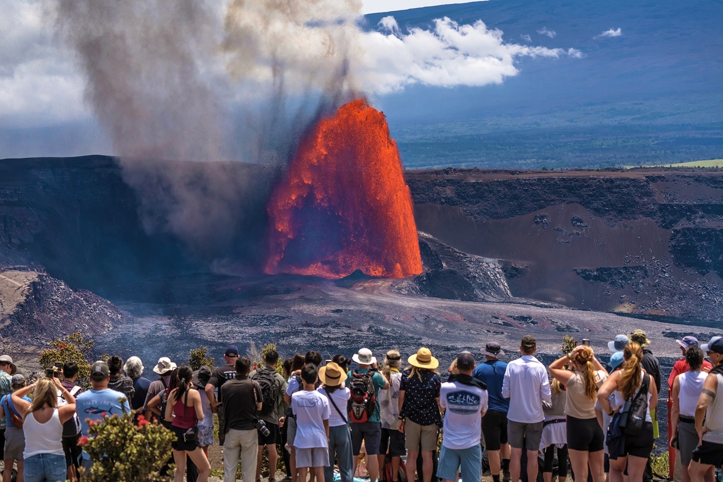 A large group of visitors observe a massive fountain of lava erupt from a volcanic vent