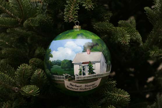 A spherical ornament painted with a scene of a small white cottage hangs from the bough of a spruce tree.