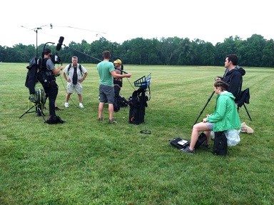 Several people on a grassy field holding video equipment and watching people being interviewed