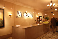 Lobby of museum. Letters MOCA are painted on wall behind marble counter. There is an orange glo to the photo. Several people are standing at the far left.