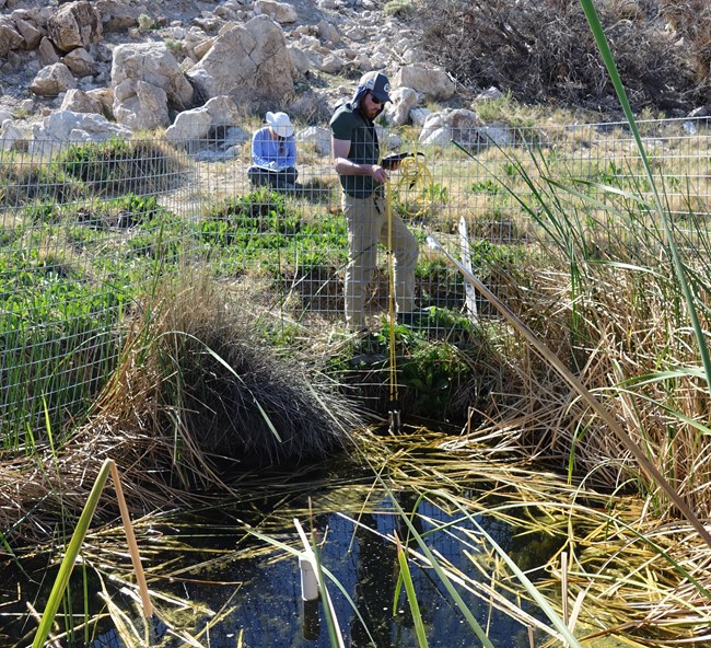 A scientist reads water quality data using a water monitoring device while another records this data on a datasheet in the background.