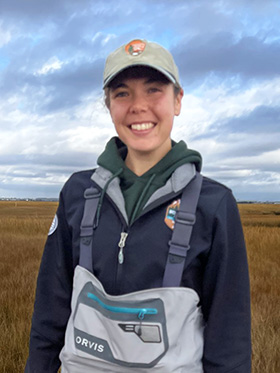 Scientist wearing hat and waders smiles at camera. She is standing in a salt marsh, with the new york city skyline behind her in the distance.