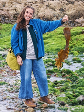 Scientist stands holding seaweed on some rocks wearing fleece and green bag,