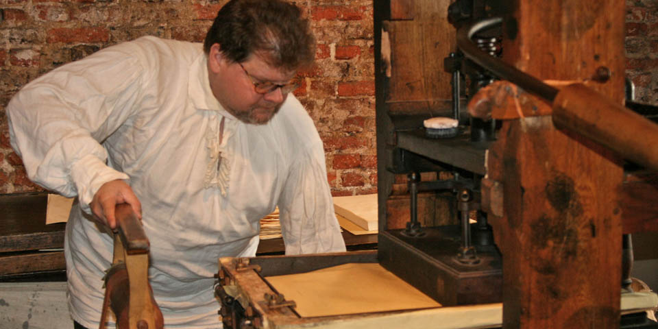 Color photo of man in 18th-century costume demonstrating the printing press.