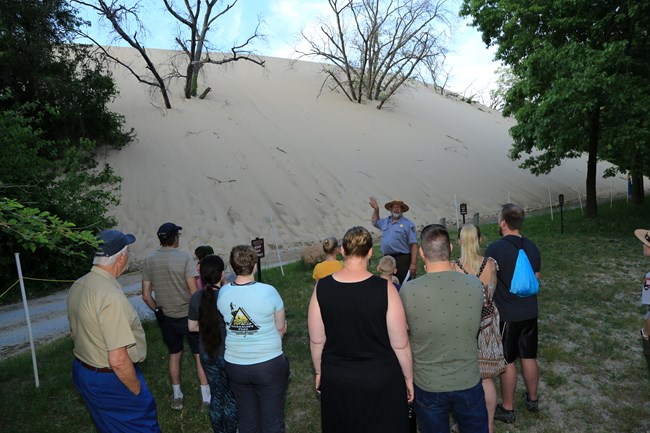 Park ranger stands at the base of the backside of a sand dune, surrounded by visitors.