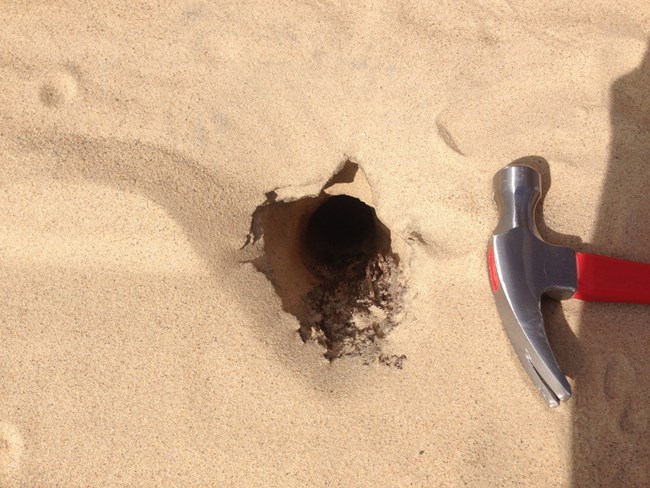 A hole on the surface of loose sand; a hammer for scale.