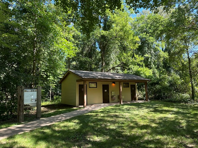 Tan building with a brown over-hanging roof surrounded by trees. A paved path leads to the front to access restroom doors and drinking fountain.