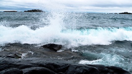 A photograph shows a wave rolling onto a rocky shoreline with an overcast sky