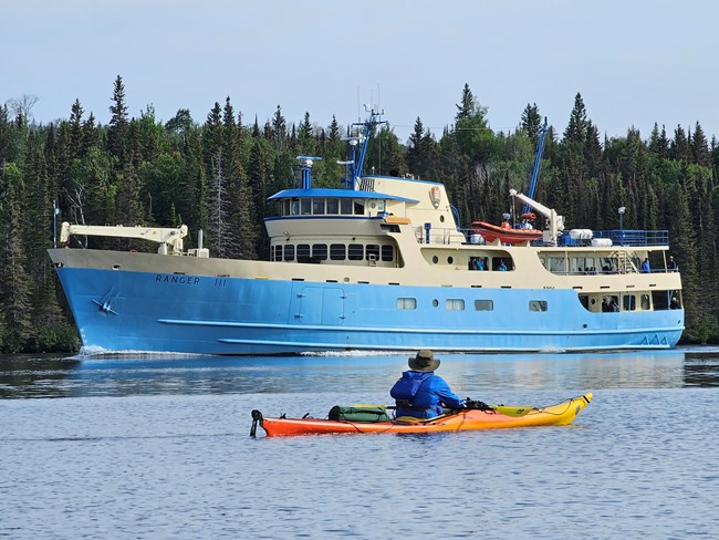 A man kayaking in front of the NPS vessel the RANGER III ferry on a sunny day.