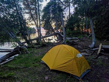 a yellow tent set up near Lake Superior with a sunset shining through the trees behind the campsite