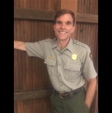 National Park Service Park Ranger in gray shirt and green pants leaning against a wood wall
