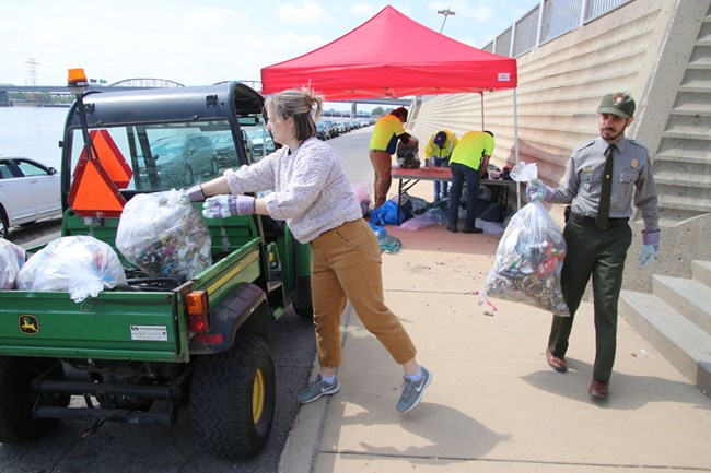 A young woman lifts a garbage bag into a small utility vehicle as a male park ranger carries another garbage bag