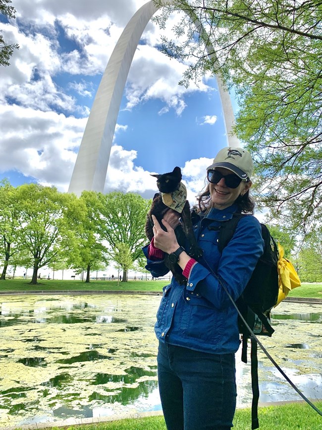 a woman holds a black cat who is wearing a bandana and a leash. The gateway arch is in the background.