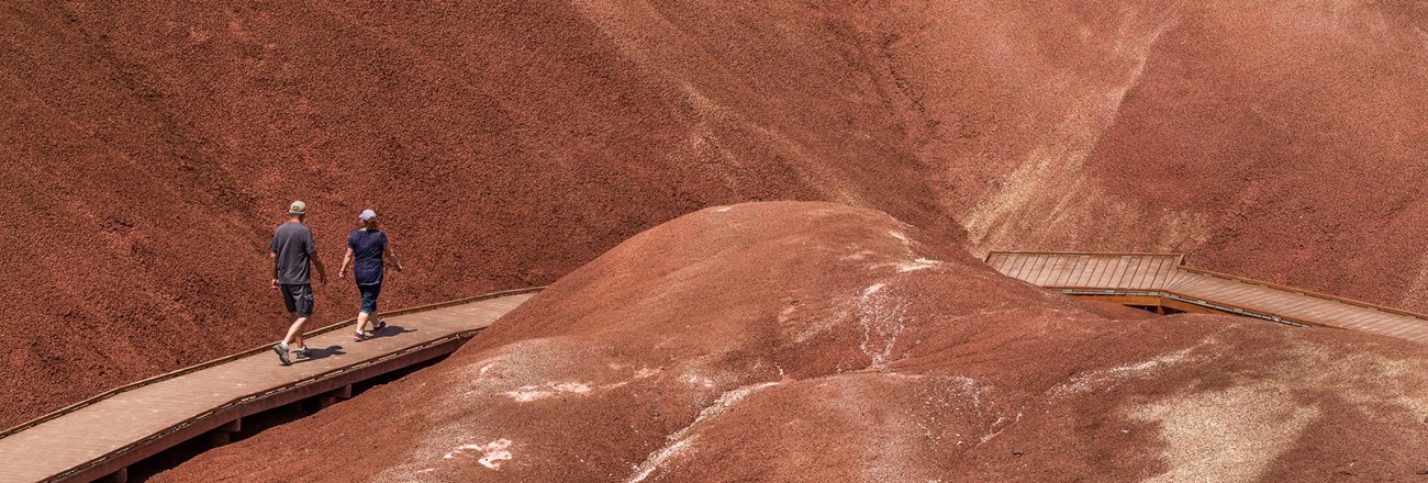 Two people walk on a boardwalk. They are surrounded by hills of deep red soil.