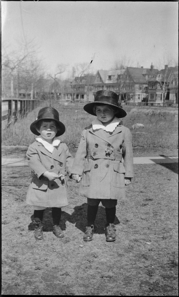 Two small boys stand outside in matching autumn clothing. The smaller boy points to his hand, being held by the bigger boy.