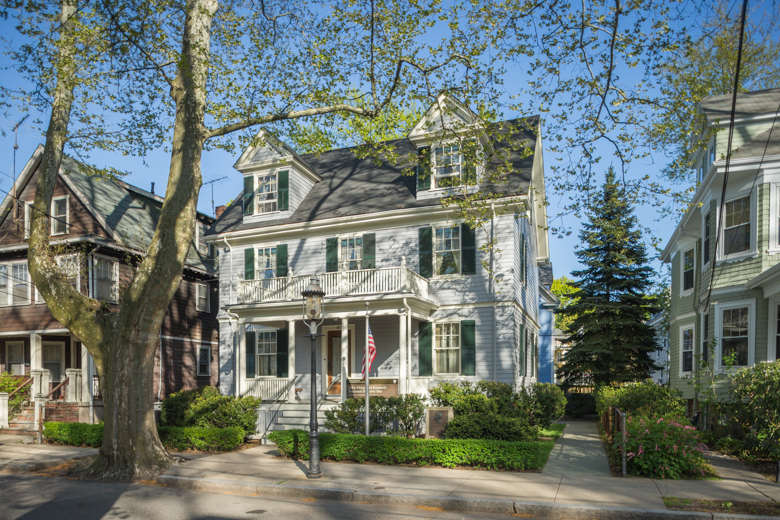 A three story grey house with dormers in its roof.  An American flag is flying on a pole in front, and a gas lamp is on the sidewalk. A large street tree with light green leaves is on the sidewalk.