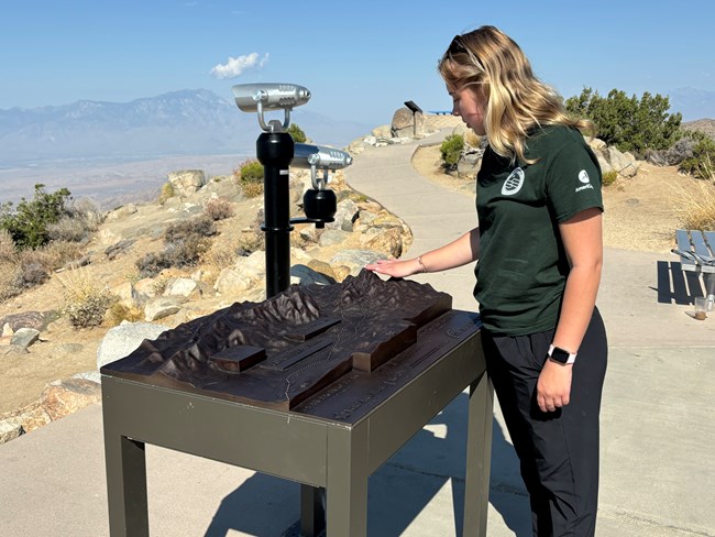 A woman touches a tactile map of mountains at a viewpoint