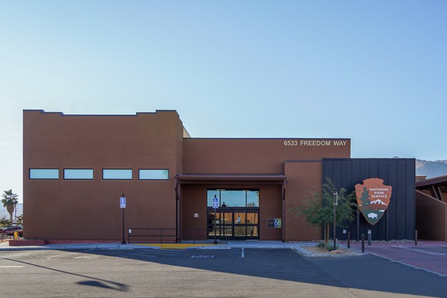 An outside view of a large brown building with a parking lot and large NPS Arrowhead