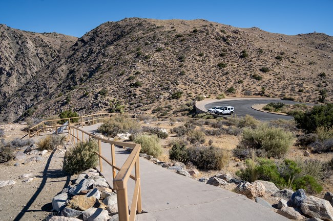 A handrail along a sidewalk on a sloped hill facing the parking lot