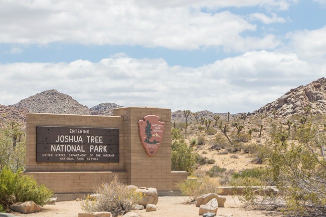 The west entrance sign with desert landscape and partly cloudy blue sky in the background.