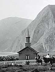A black and white image of a church building, a few people on horseback, and tall sea cliffs in the background.