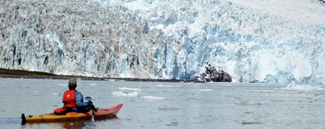 A kayaker pauses in the water near the face of a glacier.