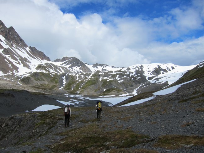 Two hikers with gear walk up towards mountains.