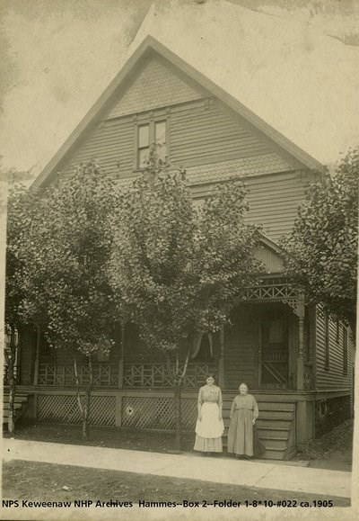 Two women with aprons stand in front of a house with trees.