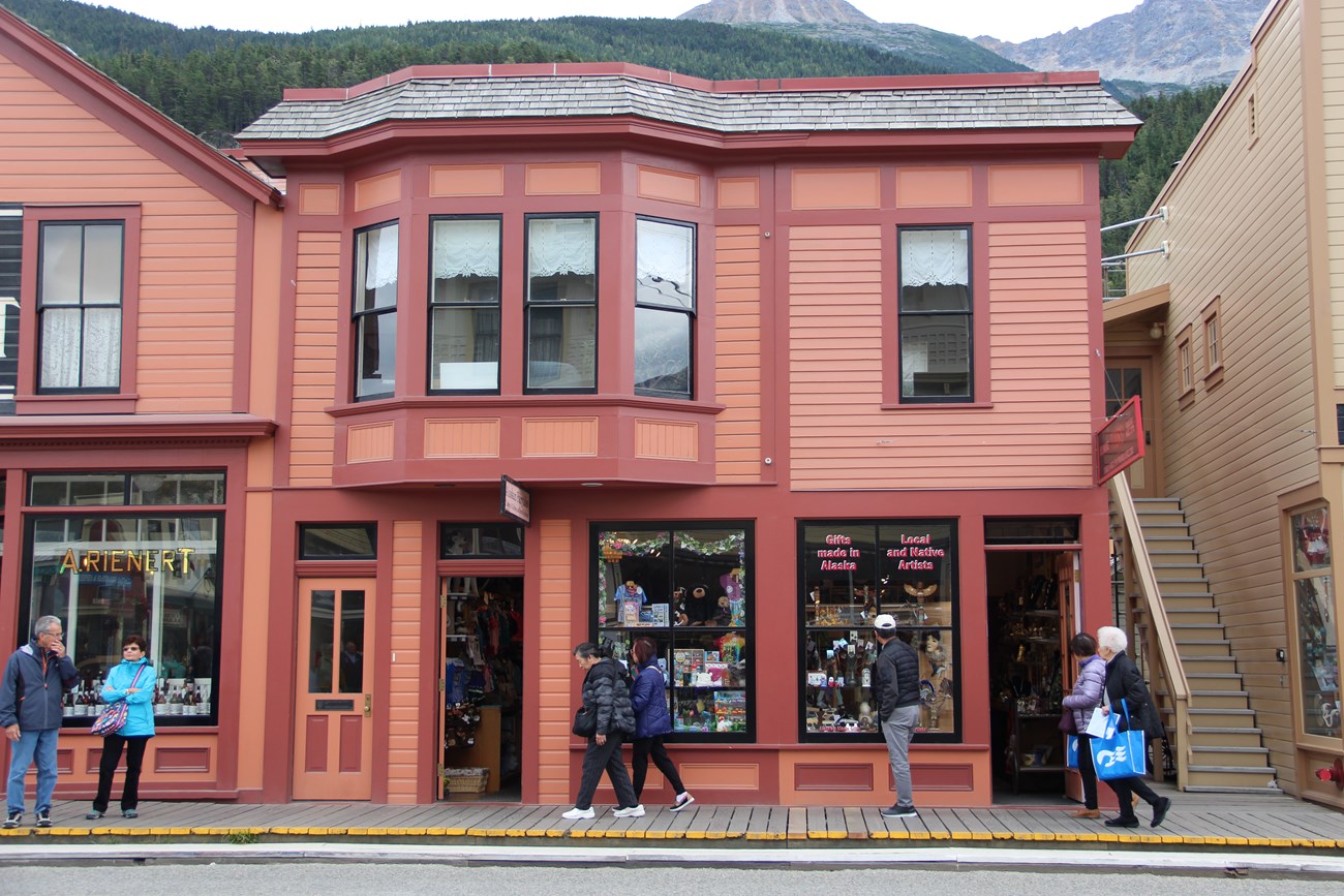 Two story pink and reddish hued Victorian style building with people walking in front
