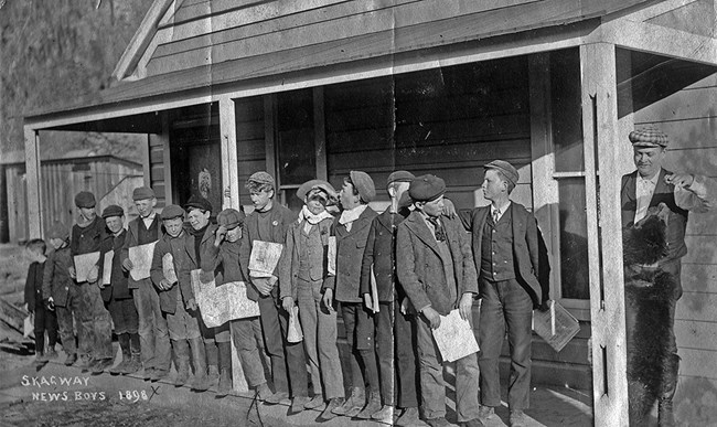 A dozen newsboys stand on a porch holding newspapers