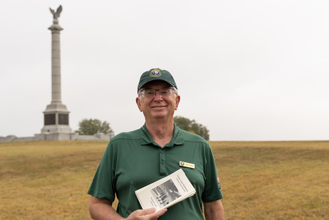 A man in a green volunteer shirt and hat holds a pamphlet in front of a tall monument in a grassy field.
