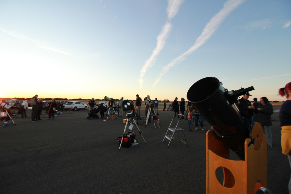 People gather around telescopes as the sun sets.