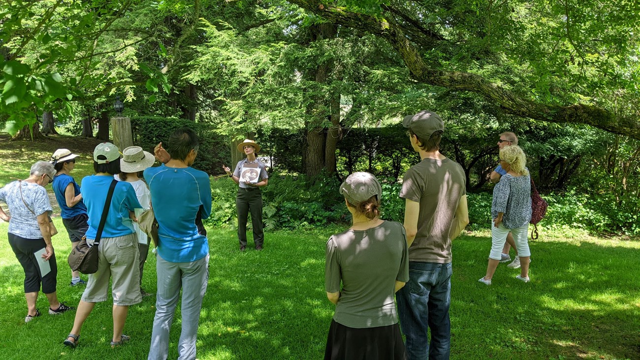 Park Ranger stands on green lawn with trees behind and talks to group of nine people