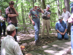 Eight people kneel on or stand next to a wooden boardwalk in the forest, discussing good ways to manage trails. NPS Photo.