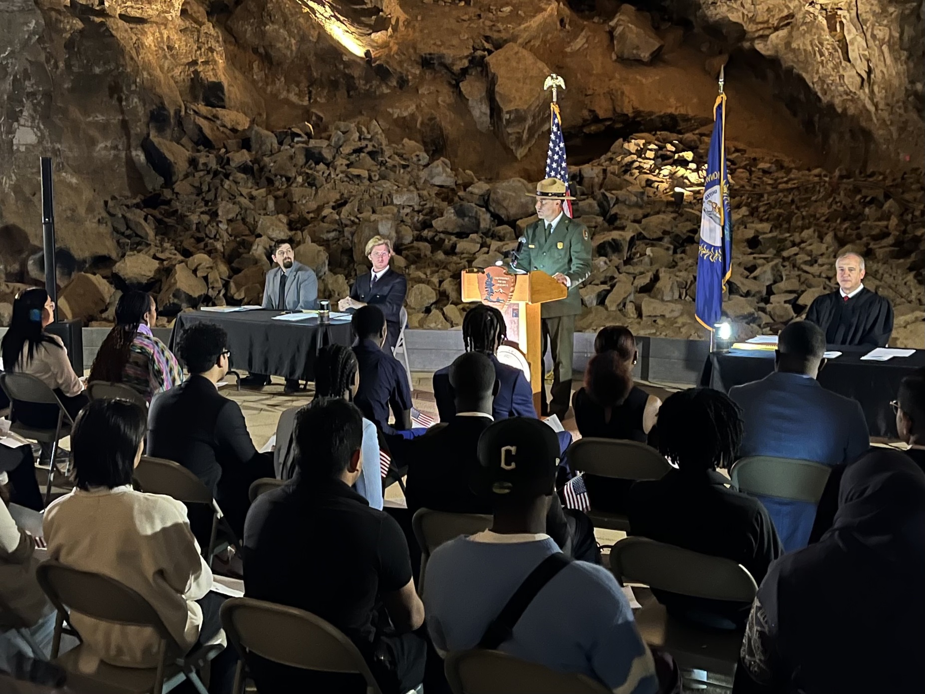 A man stands at a podium in an underground cave passageway while speaking to a group seated in front of him.