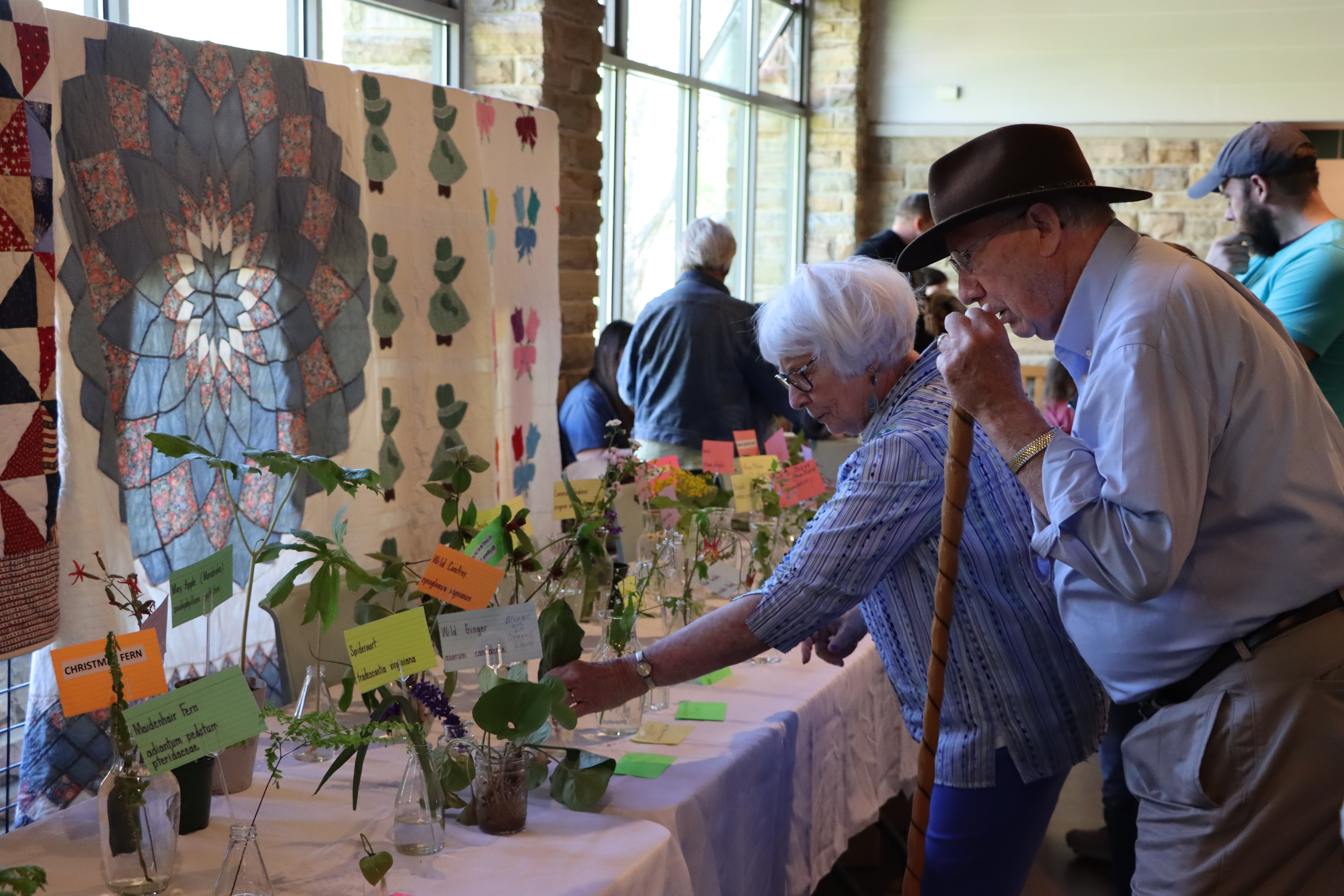 People admire colorful wildflowers on tables with quilts displayed in the background.