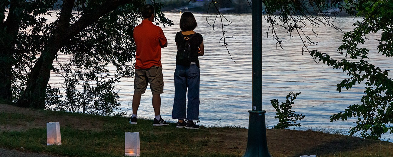 Two people in front of luminarias lining a path look at a river in twilight,