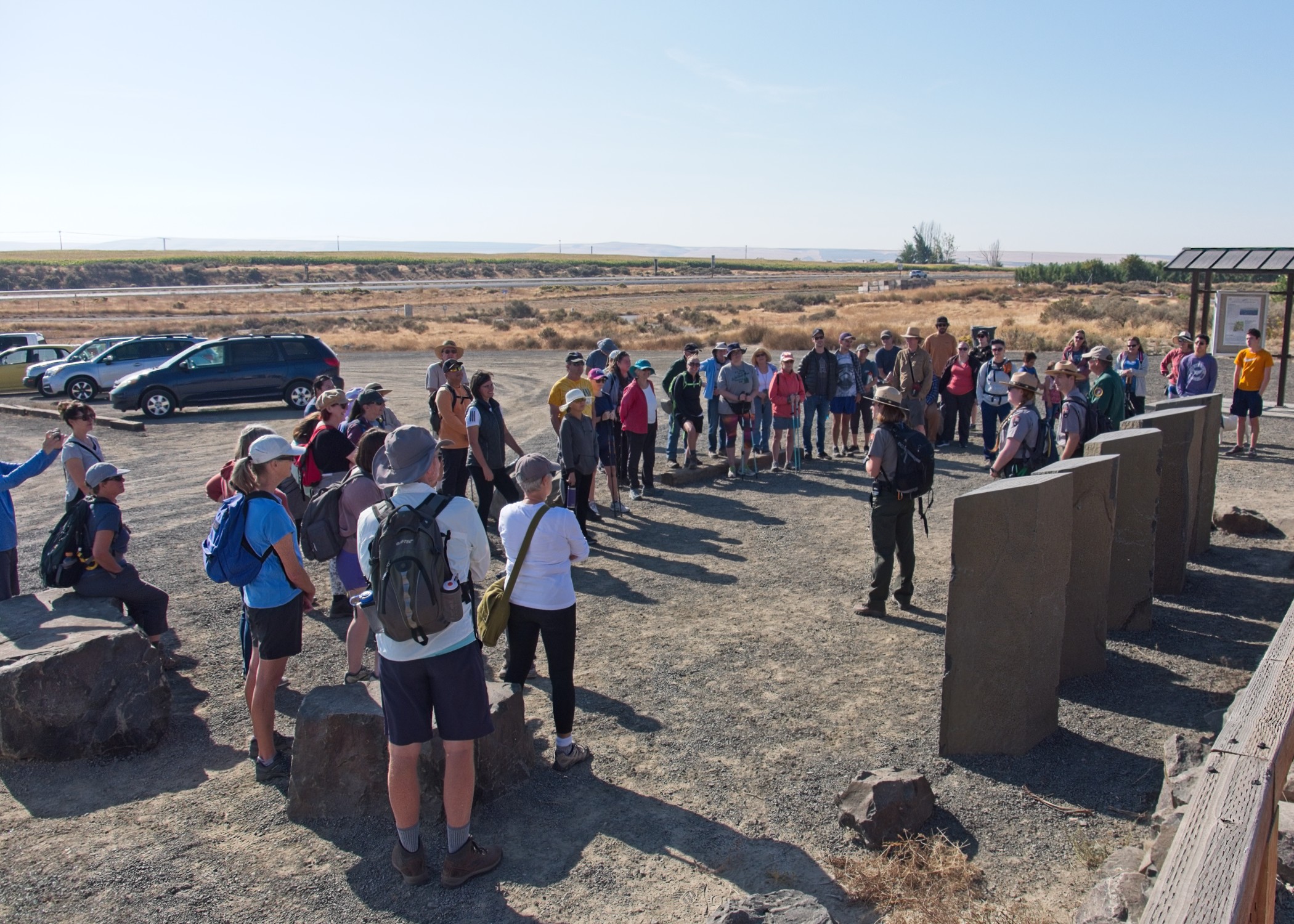 A group of people stand in a semi-circle around a ranger in uniform. Open sky and hills can be seen in the background.