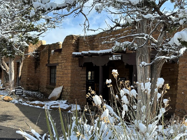 An early spring snow sits atop plants including yuccas, and pinyon and juniper trees that frame the entrance to a tan, stone masonry building with dark brown vegas. A brown sign with white letters, Museum, is hung above the covered entry.