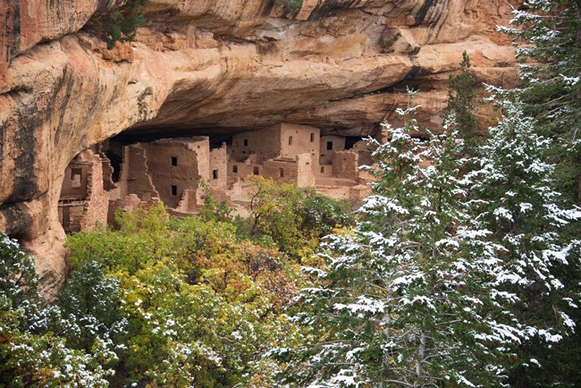 A stone masonry village sits under a sandstone alcove behind snow-covered firs