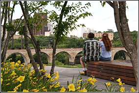 Two people sitting on a bench amidst flowers and trees overlooking a stone arch bridge.