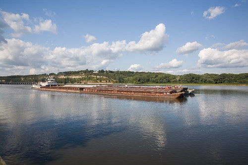 A large, white tow boat pushes a raft of barges downstream on the Mississippi River under blue sky and white, puffy clouds.