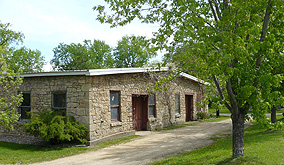 A rustic one-story, stone building sits near a path surrounded by small trees.